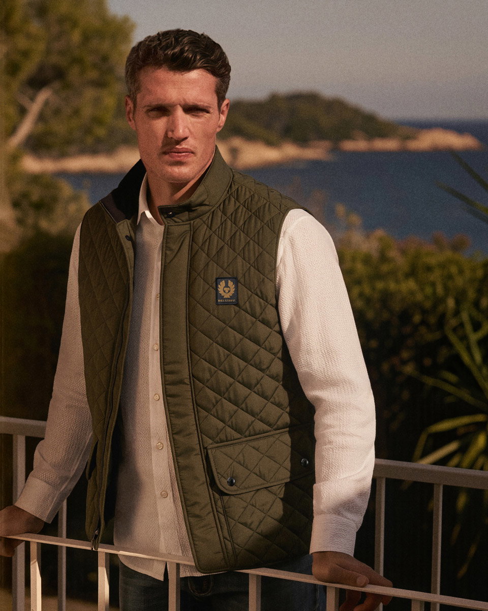 Man wearing an olive green quilted vest over a white textured shirt, standing by a balcony with a scenic ocean and greenery background.
