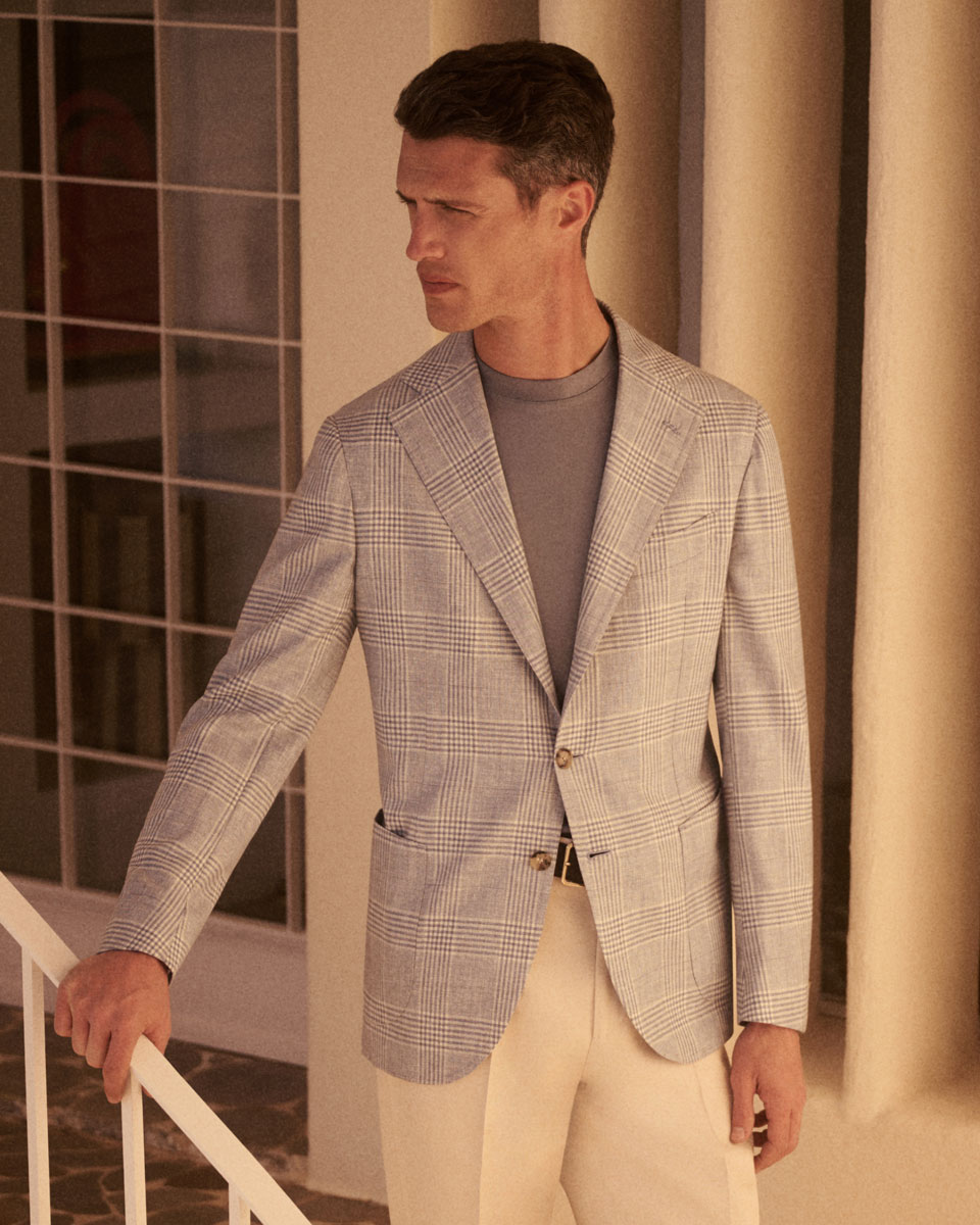 Mann trägt kariertes graues Sakko, graues T-Shirt und helle Hose, steht neben Treppengeländer vor Fenster mit weißen Säulen.
