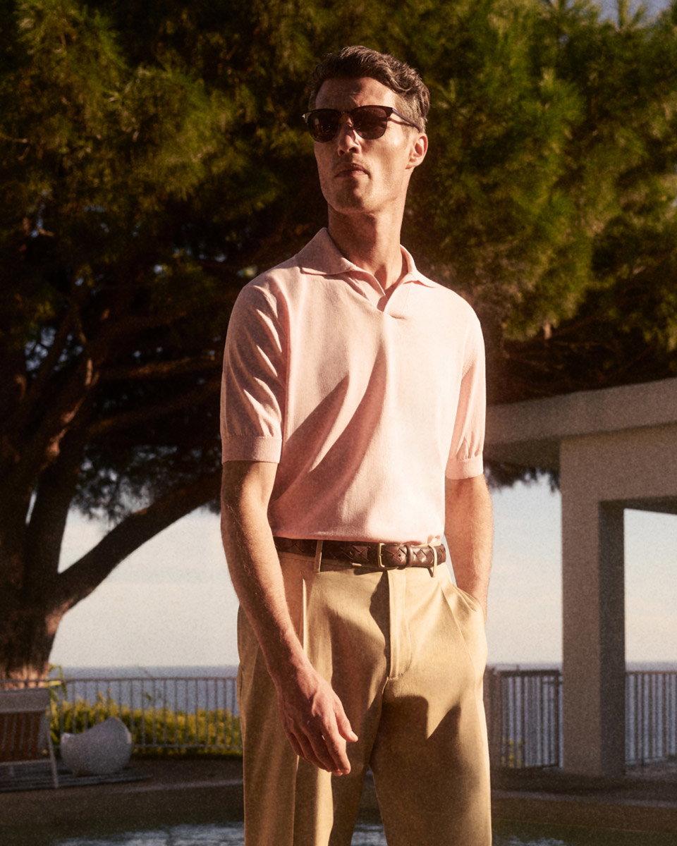 Mann mit brauner Sonnenbrille, rosa Polohemd und beiger Hose steht vor Bäumen und Meer in sonniger Außenanlage.