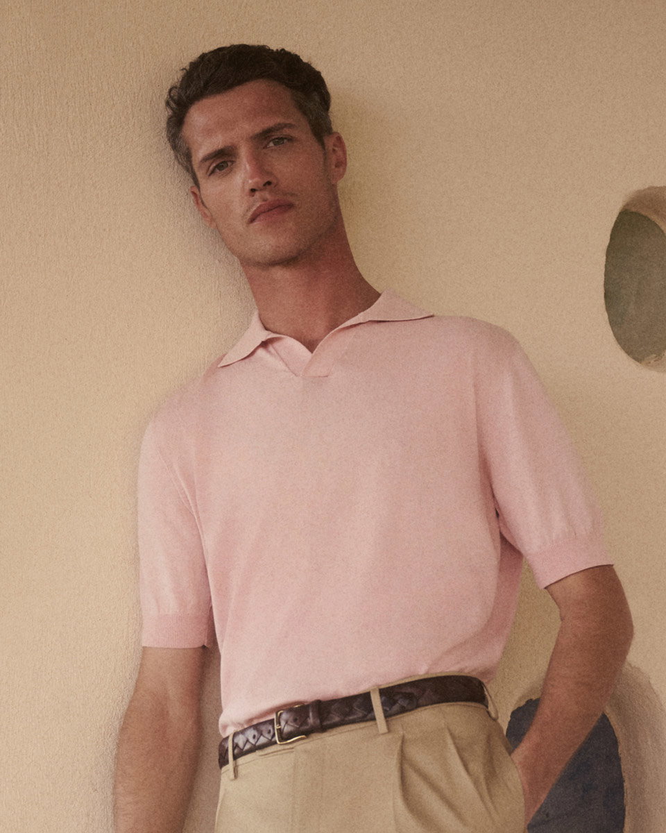 Mann lehnt an beiger Wand, trägt hellrosa Kurzarm-Polo, beigefarbene Hose und braunen geflochtenen Ledergürtel.