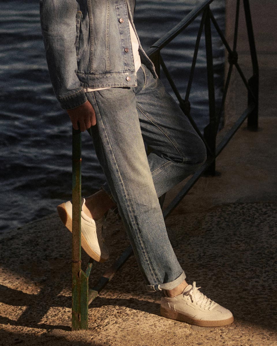 Mann lehnt am Geländer am Wasser, trägt blaue Jeans, Jeansjacke und weiße Sneaker mit brauner Sohle.