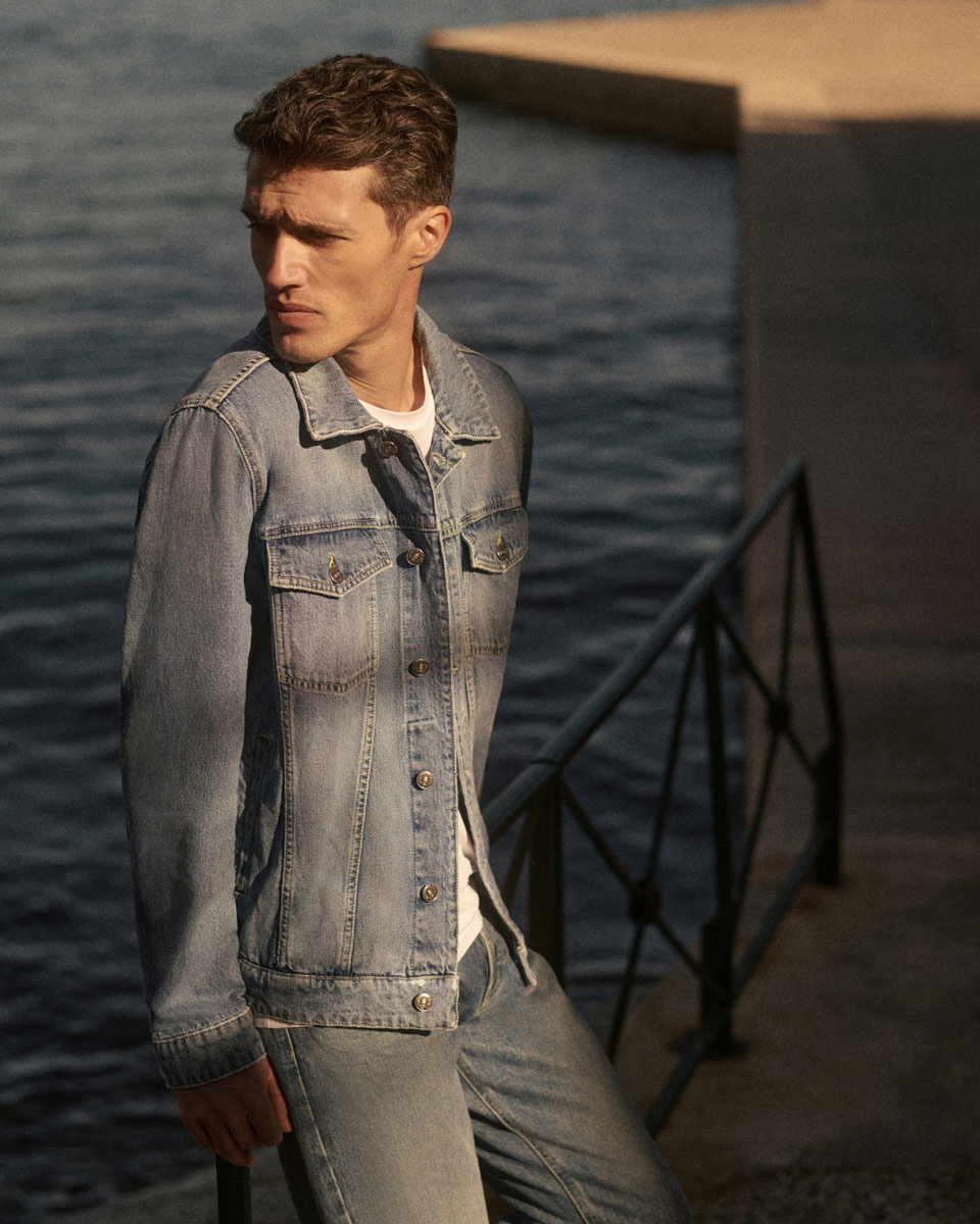 Mann trägt helle Jeansjacke und Jeanshose, steht am Wasser mit Geländer, schaut nach links, Sonnenlicht fällt auf ihn.