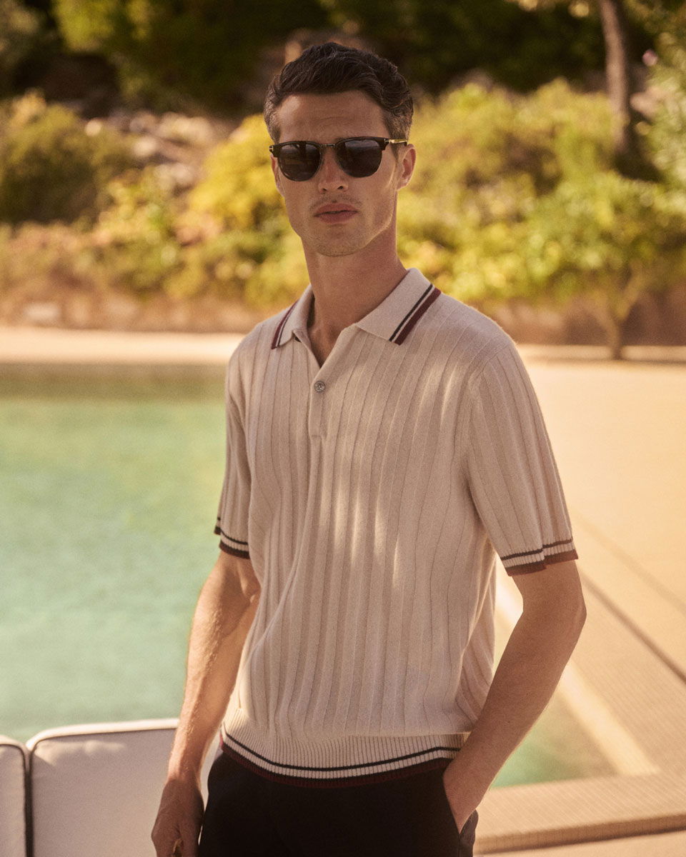 Junger Mann mit dunklen Haaren, Sonnenbrille und cremefarbenem Poloshirt steht entspannt neben einem Pool im Freien.