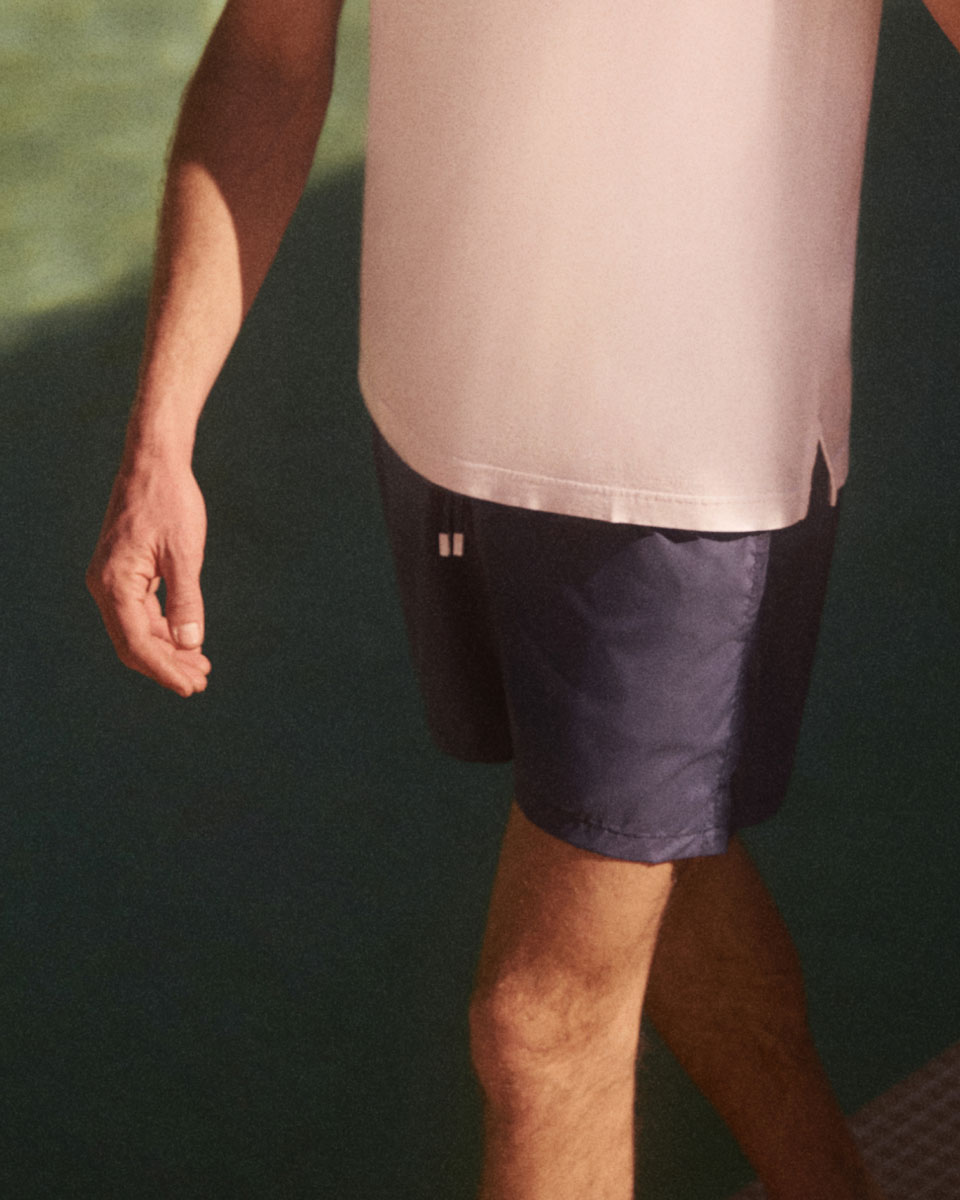 Mann trägt weißes T-Shirt und dunkelblaue Shorts, geht am Wasser entlang, sichtbarer Arm und Beine im Sonnenlicht.