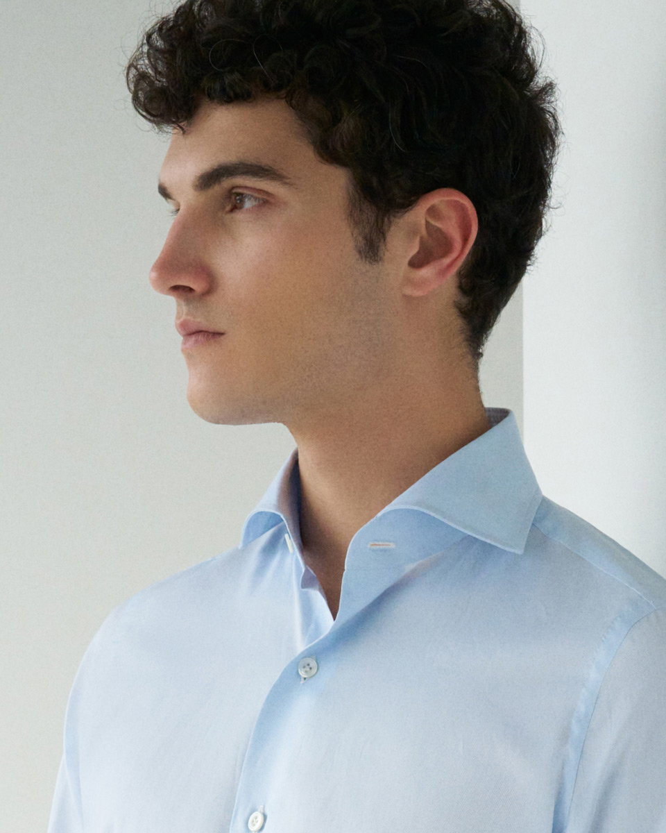 Junger Mann mit lockigem dunklem Haar trägt hellblaues Hemd mit offenem Kragen, Blick nach links, neutraler Hintergrund.