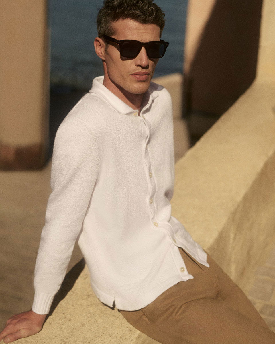 Mann mit brauner Hose und weißem Strickhemd trägt schwarze Sonnenbrille, sitzt draußen auf gelbem Steinmauer, sonnig.