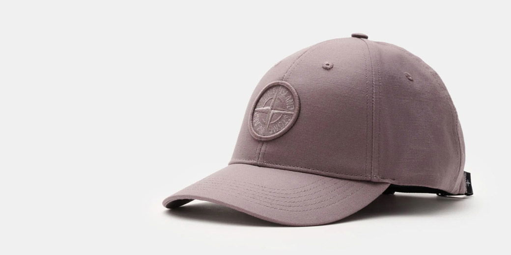 Grayish-purple baseball cap with stitched circular compass logo on front, curved brim, and adjustable strap at back.