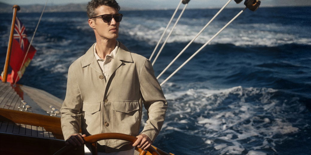 Mann mit Sonnenbrille steuert Segelboot bei sonnigem Wetter, trägt beige Jacke, im Hintergrund blaues, bewegtes Meer.