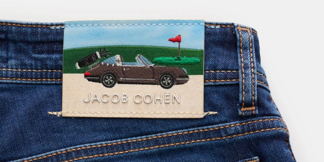Jeans mit brauner Naht und einem Markenlabel aus Stoff, das einen Cabrio-Sportwagen, Golfloch mit Fahne und den Schriftzug „JACOB COHËN“ zeigt.