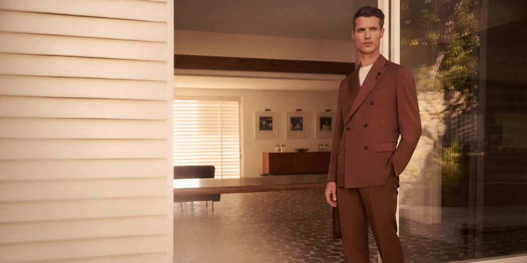 A man in a brown double-breasted suit with a white shirt stands in a modern room with a wooden table and framed pictures.