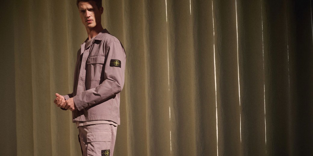 Mann trägt hellviolettes Stone Island Outfit mit Jacke und Hose vor beigen Vorhängen, Licht fällt schräg von rechts.
