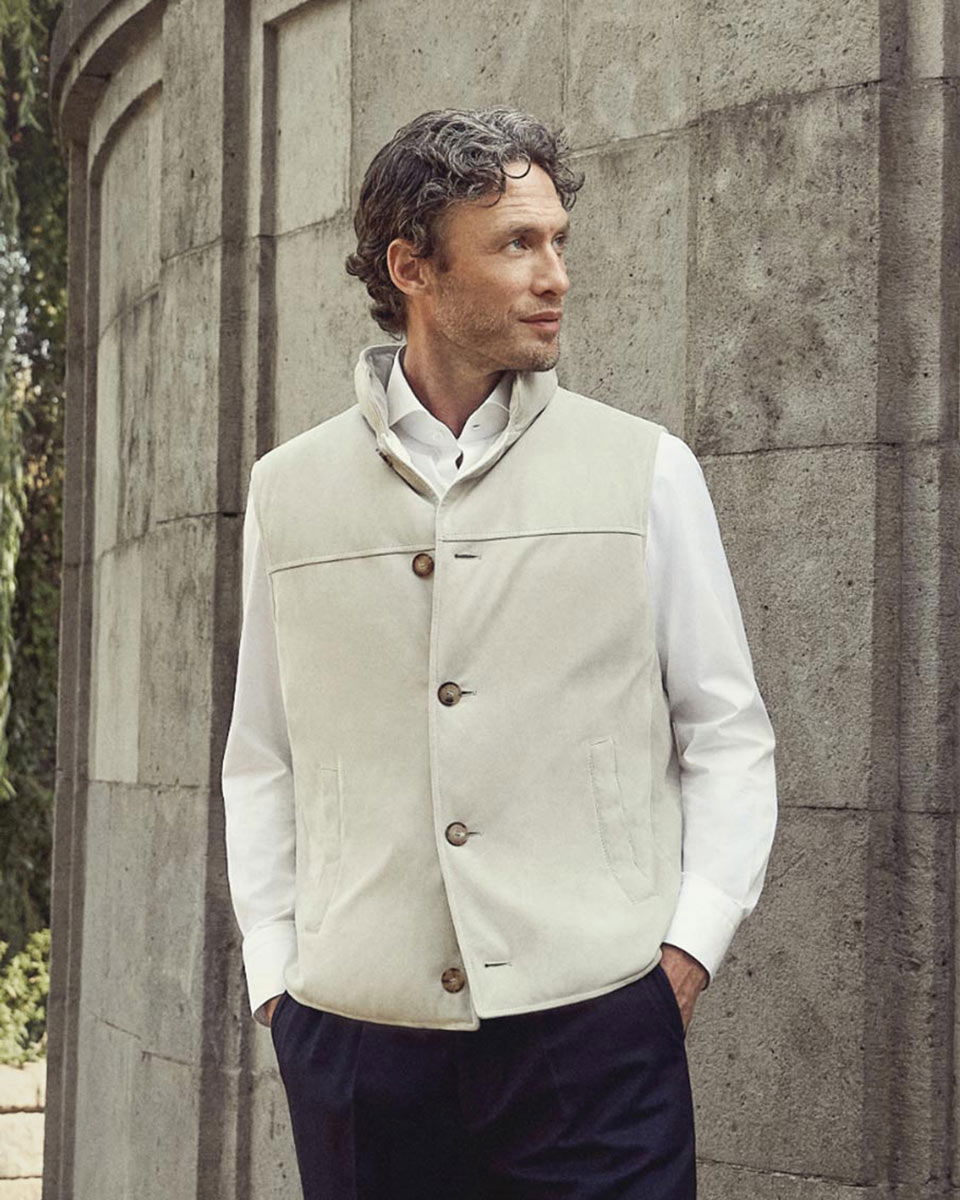 Mann mit weißem Hemd und beiger Weste lehnt an grauer Steinwand und schaut nach rechts, Hände in Hosentaschen.