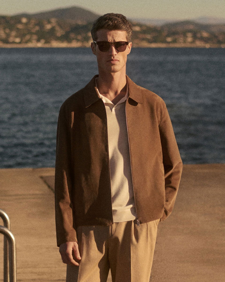 Man wearing brown leather jacket, white polo shirt, and beige pants standing near water with mountains in the background.