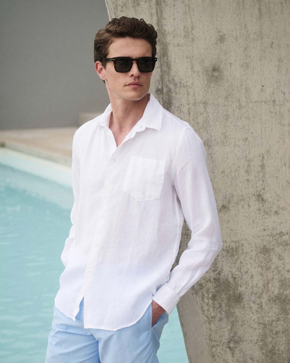 Young man wearing black sunglasses, a white linen button-up shirt, and light blue pants, leaning against a concrete wall by a pool.