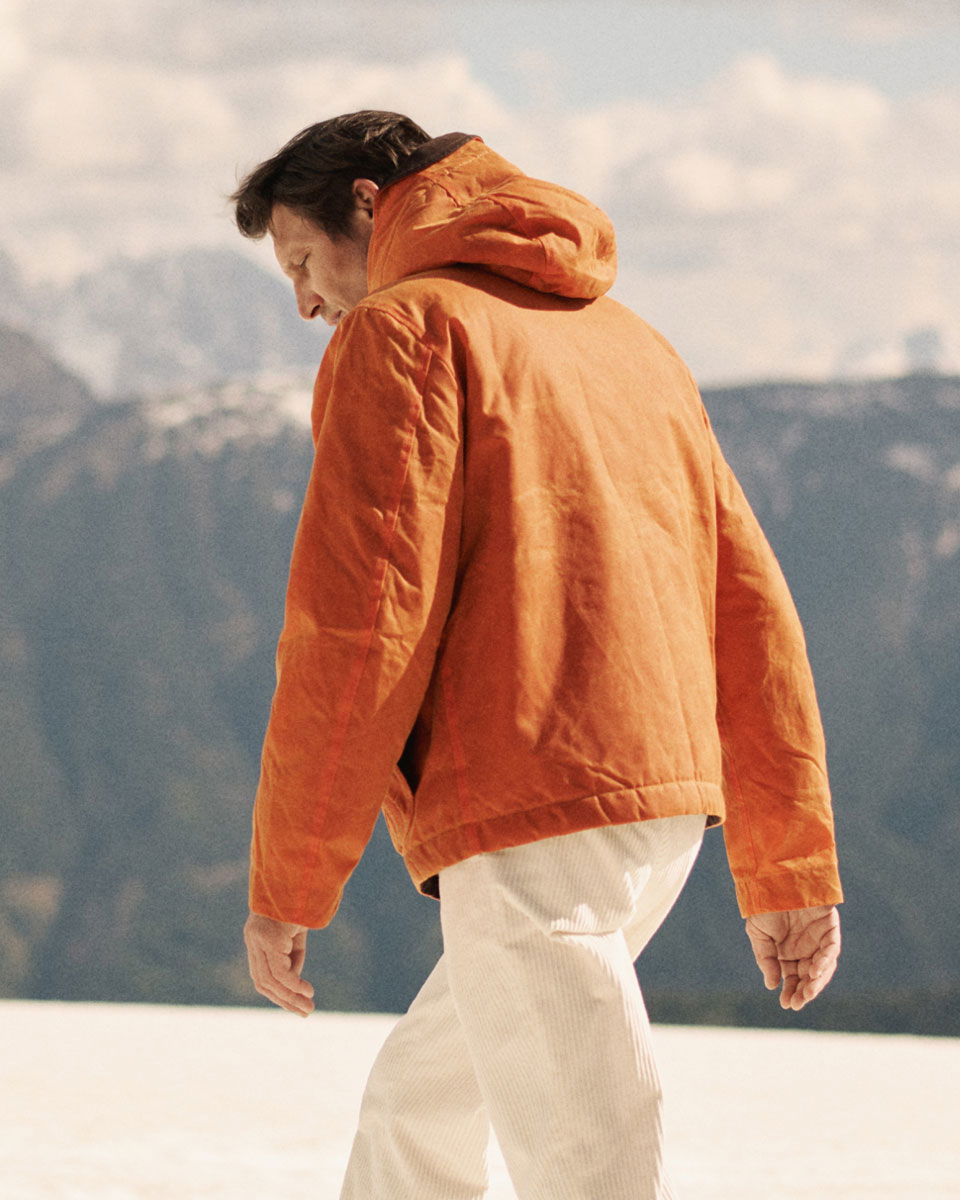 Man wearing an orange hooded jacket and white pants walking outdoors with mountains and cloudy sky in the background.