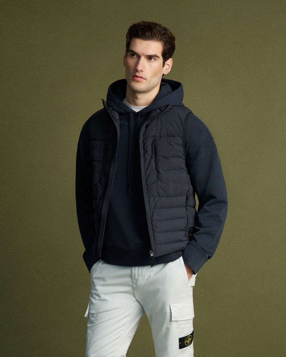 Young man wearing a black padded vest over a navy hoodie and white cargo pants with hands in pockets against olive background.