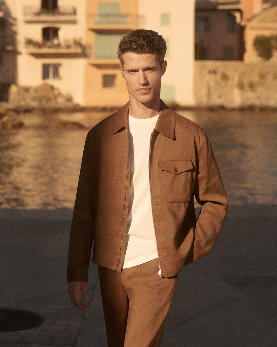 Man wearing brown overshirt and matching pants with a white t-shirt, standing outdoors near water and buildings in warm light.