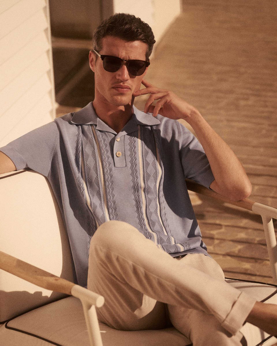 Mann mit dunkler Sonnenbrille, hellblauem Strick-Poloshirt und beiger Hose sitzt entspannt auf weißem Stuhl im Sonnenlicht.