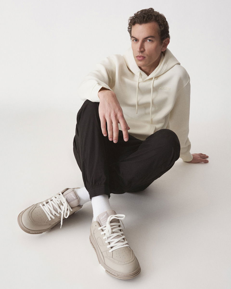 Mann sitzt auf Boden, trägt cremefarbenen Hoodie, schwarze Hose, weiße Socken und beige Sneaker mit weißen Schnürsenkeln.