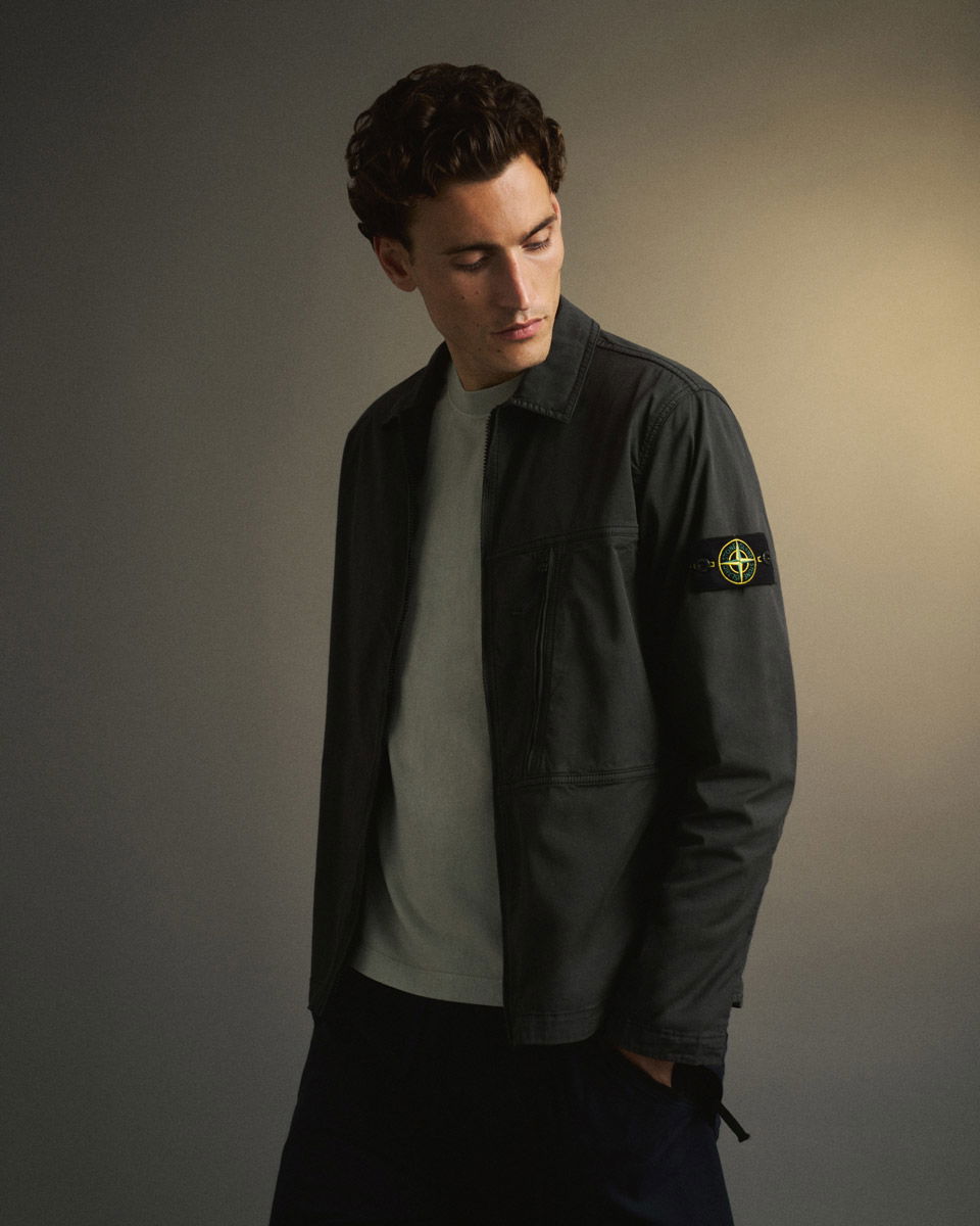 Junger Mann mit dunklem, lockigem Haar trägt schwarze Stone Island Jacke über grauem Shirt, schaut nach unten.