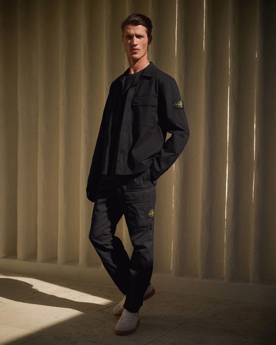 Mann trägt schwarze Stone Island Jacke und Hose mit Logo-Patches, weiße Sneakers, steht vor beiger Wand mit Lichtstreifen.