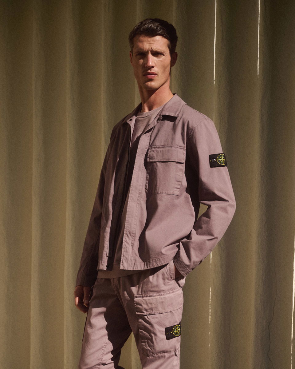 A man wearing a mauve Stone Island jacket and matching cargo pants with logo patches, standing against a beige curtain.