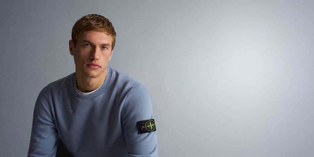 Mann mit kurzem blondem Haar trägt hellblauen Stone Island Pullover mit Logo-Patch am linken Oberarm vor grauem Hintergrund.