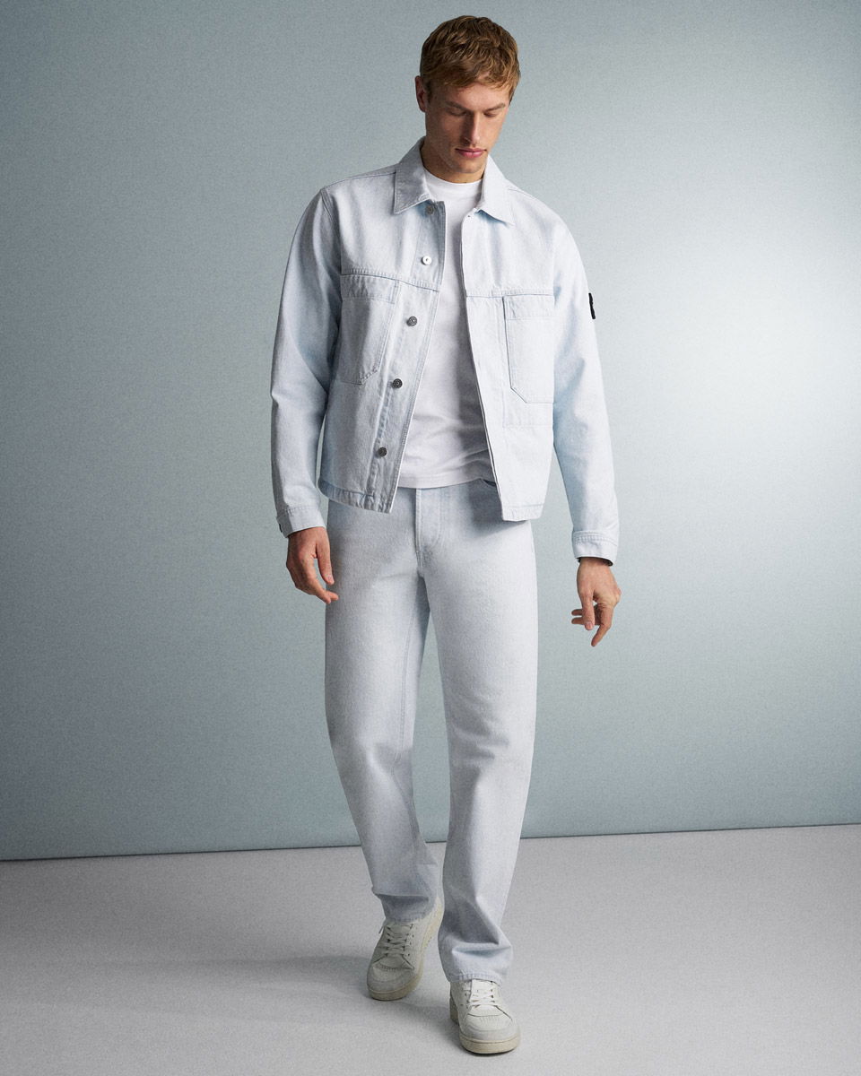 Männliches Modell trägt hellblaue Stone Island Denim-Jacke, weiße T-Shirt, helle Jeans und weiße Sneakers vor grauem Hintergrund.