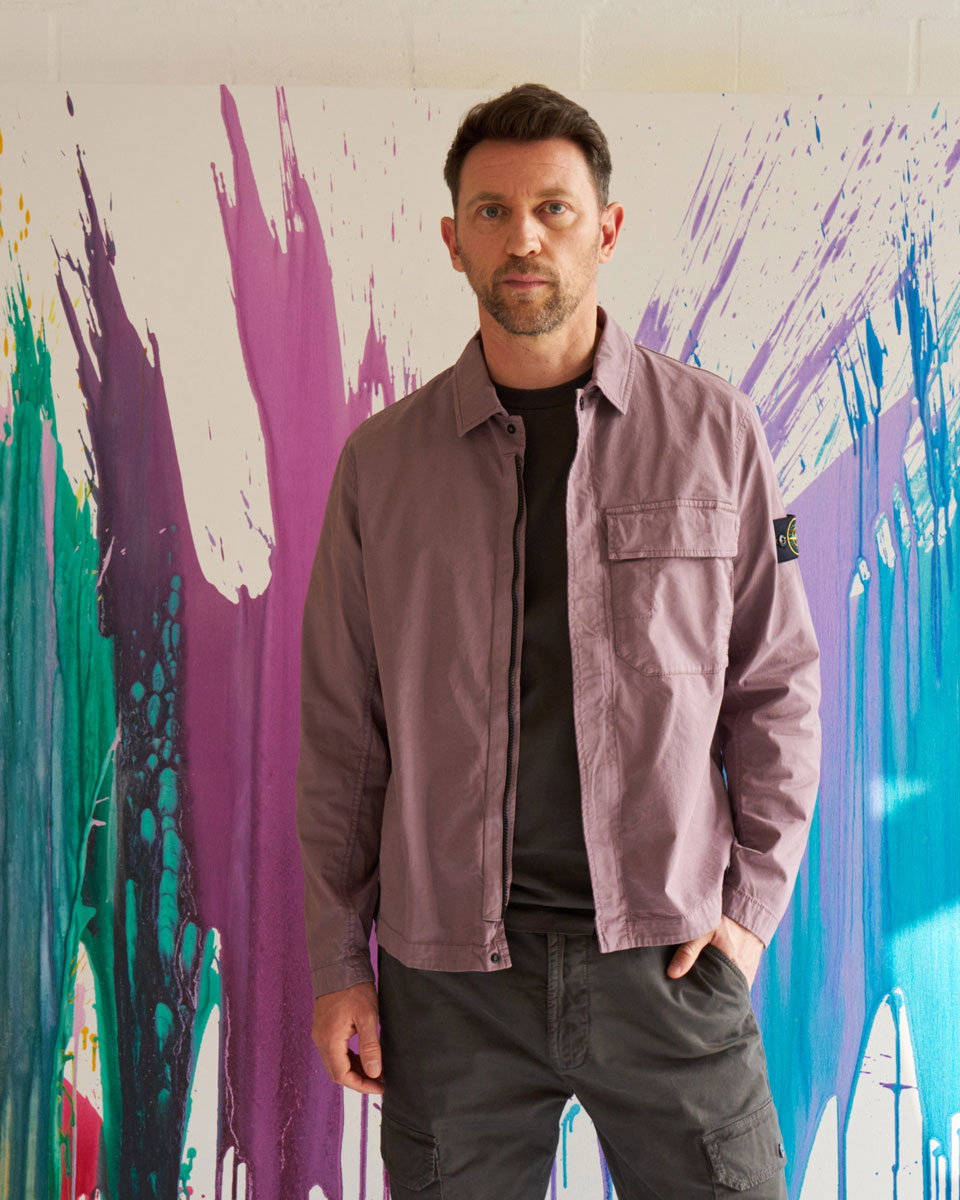 Mann mit brauner Jacke und schwarzem T-Shirt vor bunter, abstrakter, farbiger Wand, Hände in Hosentaschen.