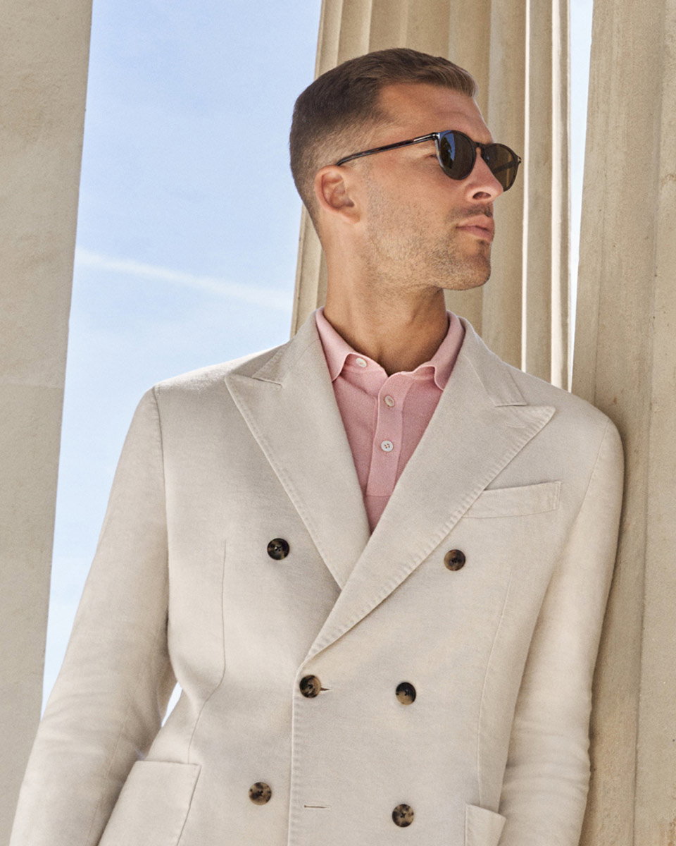 Mann mit kurzen Haaren und Sonnenbrille trägt helles, doppelt geknöpftes Sakko über rosa Hemd, steht vor Säulen.