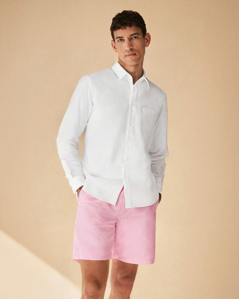 Mann trägt weißes Hemd und rosa Shorts, steht vor beige Hintergrund, Hände in Taschen, neutraler Gesichtsausdruck.