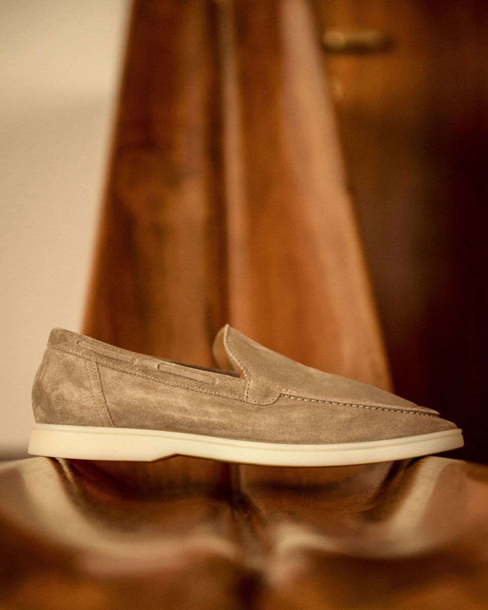 Beige suede loafer shoe with white sole placed on a polished wooden surface with warm brown background.