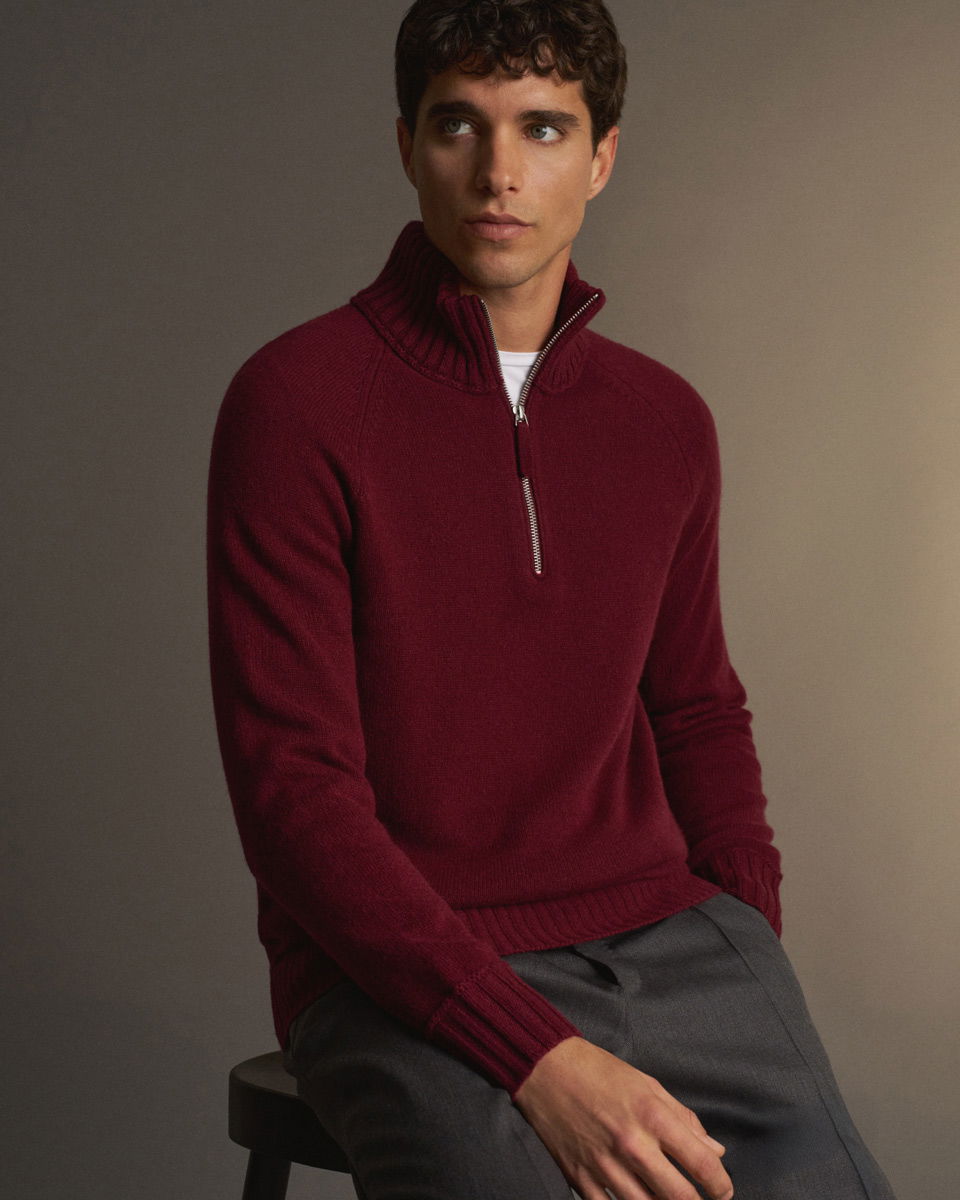 Junger Mann sitzt auf Hocker, trägt bordeauxroten Strickpullover mit halbem Reißverschluss und graue Hose vor beige Hintergrund.