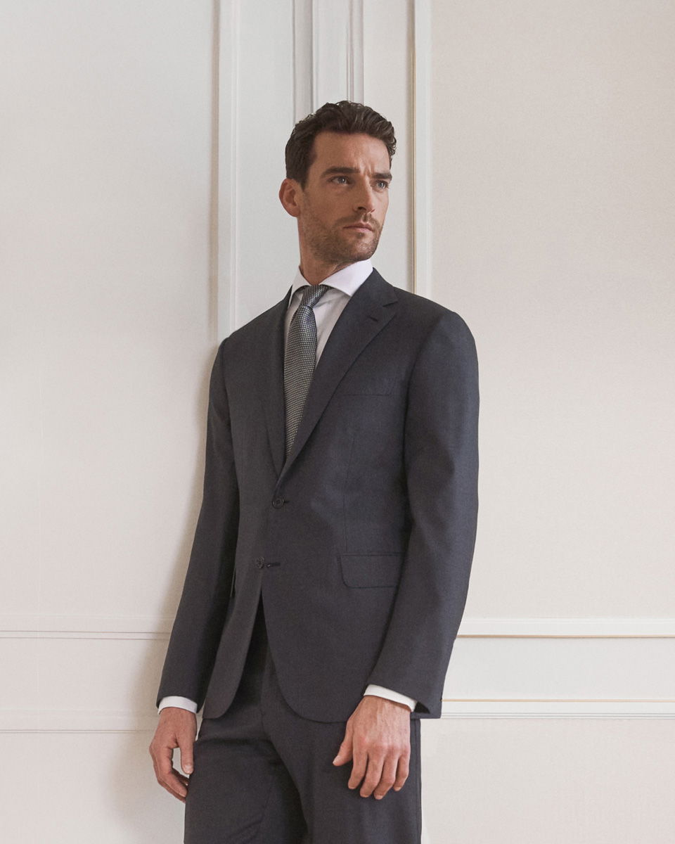 Eleganter Mann mit dunklem Anzug, weißem Hemd und gemusterter Krawatte steht vor weißer Wand im festlichen Outfit.