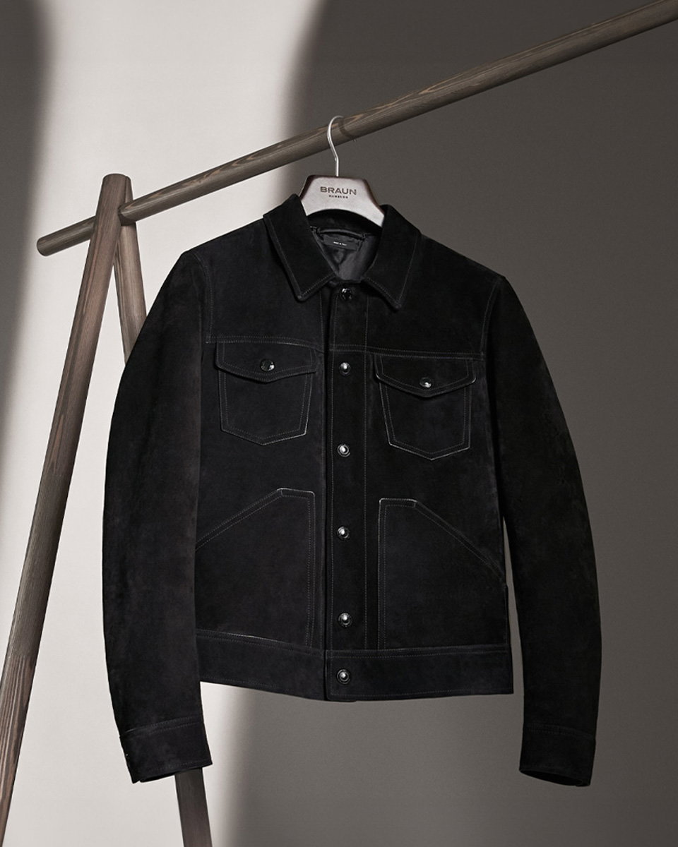 Schwarze Wildlederjacke mit Knöpfen und Taschen, hängend an einem Holzkleiderständer vor hell-dunklem Hintergrund.