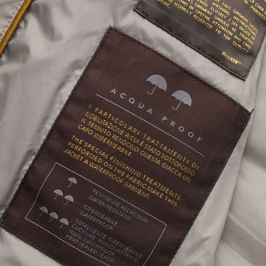 Innenansicht einer Jacke mit braunem Label „ASQUA PROOF“ und Text zu wasserdichten, spezialbehandelten Stoffeigenschaften.