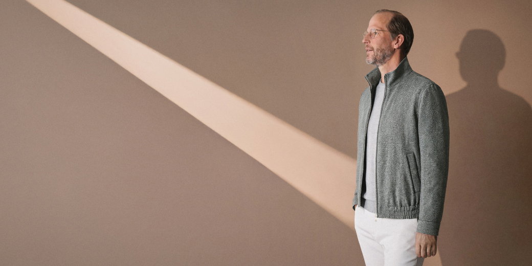 Mann mit Brille und grauer Jacke steht vor beige Wand, Sonnenstrahl fällt schräg auf die Wand, Schatten sichtbar.