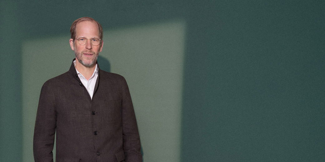Middle-aged man with glasses and beard wearing a dark brown jacket and white shirt standing against a green wall.