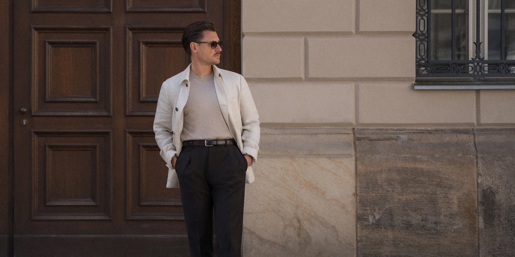 Man wearing sunglasses, light beige jacket, gray shirt, and black pants standing with hands in pockets by wooden door and stone wall.