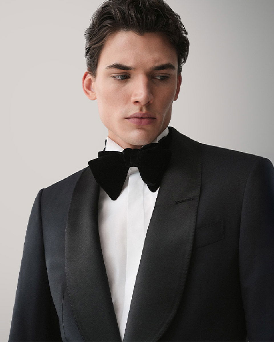 Junger Mann in elegantem schwarzen Smoking mit weißem Hemd und schwarzer Fliege vor neutralem Hintergrund, Blick nach links.