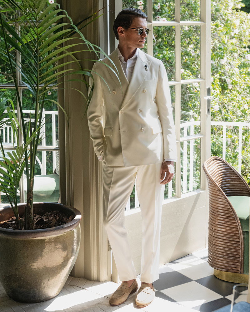 Mann in weißem Doppelreiher-Anzug und Sonnenbrille lehnt an Glastür, daneben große Topfpflanze, helles Tageslicht.