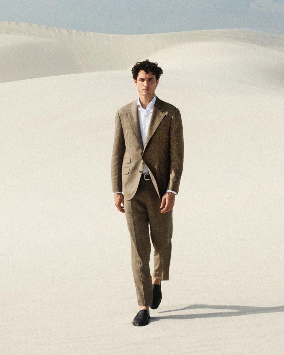 Mann im braunen Anzug mit weißem Hemd und schwarzen Schuhen läuft über helle Sanddüne unter blauem Himmel.