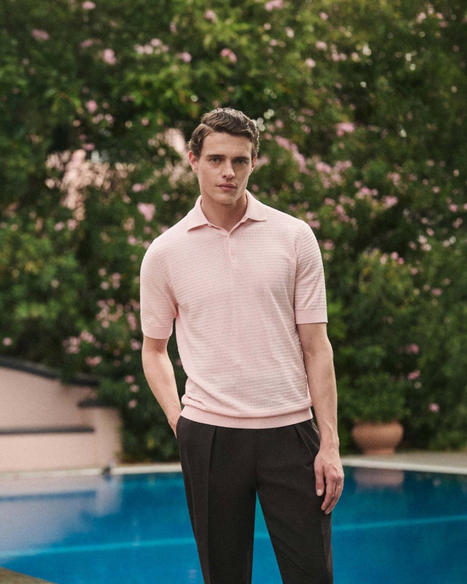 Junger Mann mit rosa kurzärmeligem Poloshirt und dunkler Hose steht vor einem blühenden Garten und blauem Pool.