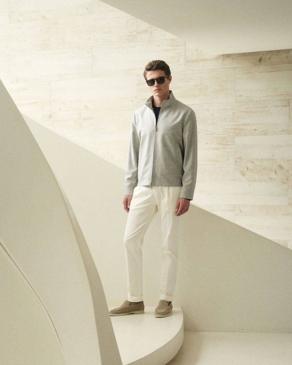 Mann mit Sonnenbrille trägt graue Jacke, weiße Hose und beige Schuhe, steht vor hellen, minimalistischen Wänden und Treppe.