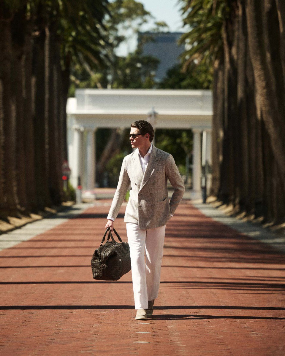 Mann in hellem Anzug und Sonnenbrille läuft auf von Palmen gesäumtem roten Ziegelweg, trägt schwarze Reisetasche.