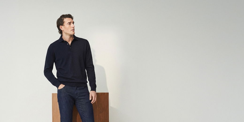 Mann mit dunkelblauem Pullover und Jeans lehnt an Holzplatte vor hellgrauer Wand und schaut nach links.