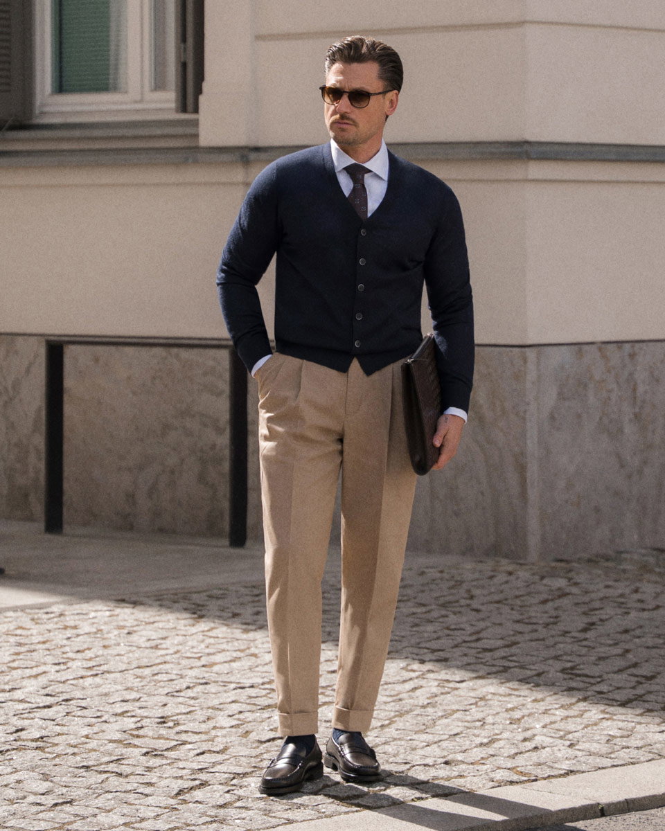 Man wearing sunglasses, navy cardigan over white shirt with tie, beige trousers, black loafers, holding brown leather portfolio.