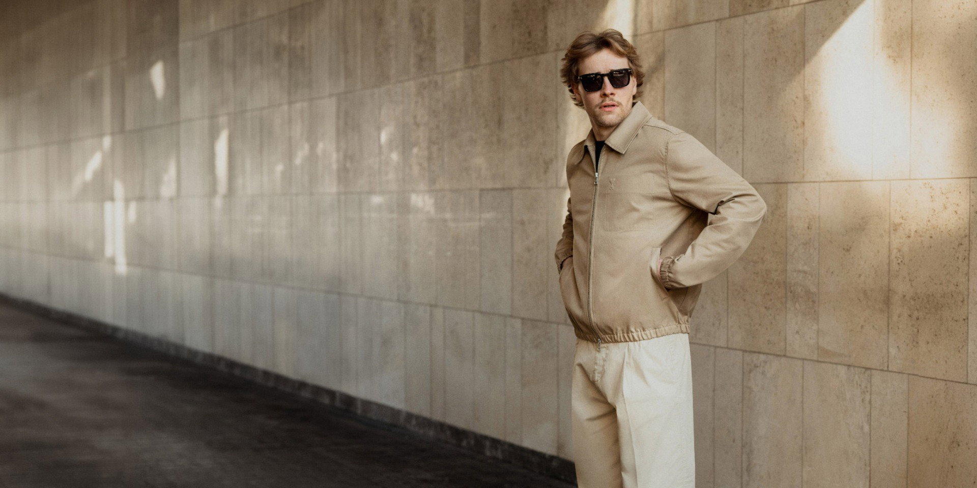 Man wearing sunglasses, beige jacket, and light pants standing with hands in pockets against a tiled wall with sunlight patches.