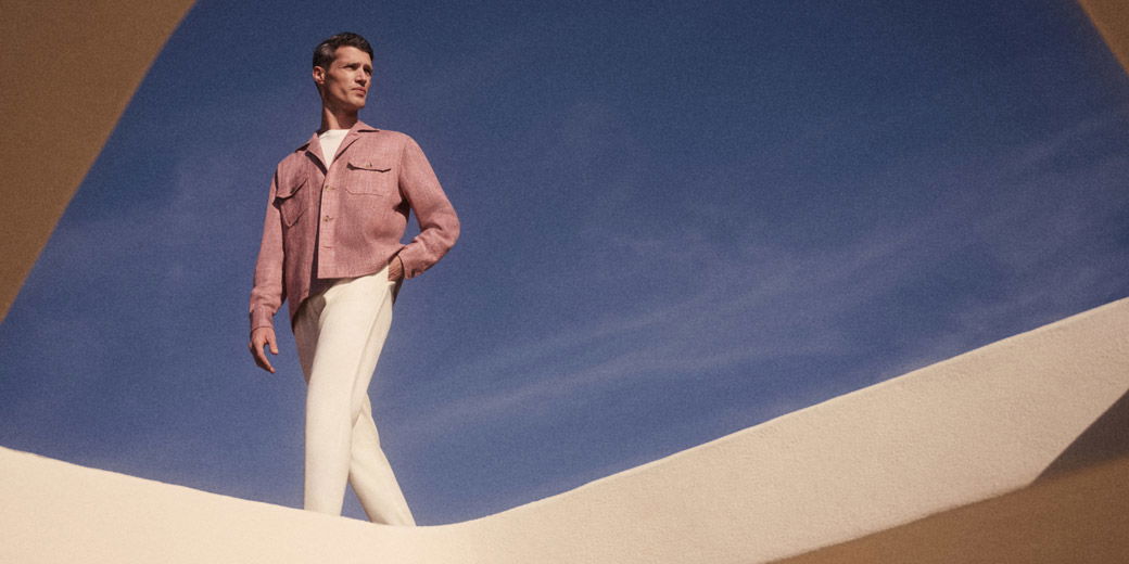 Man wearing a pink jacket and white pants standing against a clear blue sky, framed by beige architectural elements.