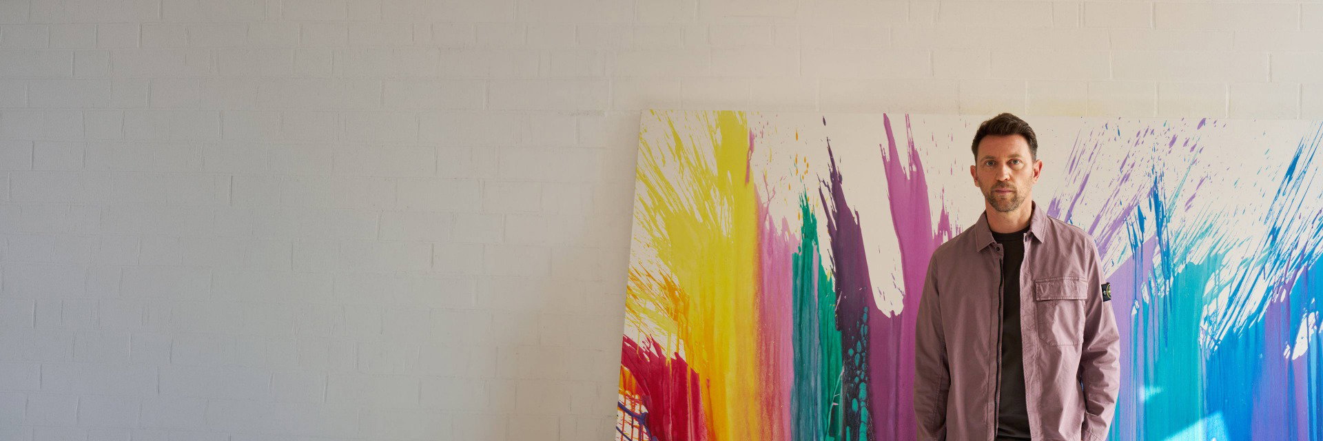 Mann steht vor großer Leinwand mit bunten Farbspritzern, trägt schwarze Hose und mauvefarbene Jacke vor weißer Wand. Mann steht vor großer Leinwand mit bunten Farbspritzern, trägt schwarze Hose und mauvefarbene Jacke vor weißer Wand.